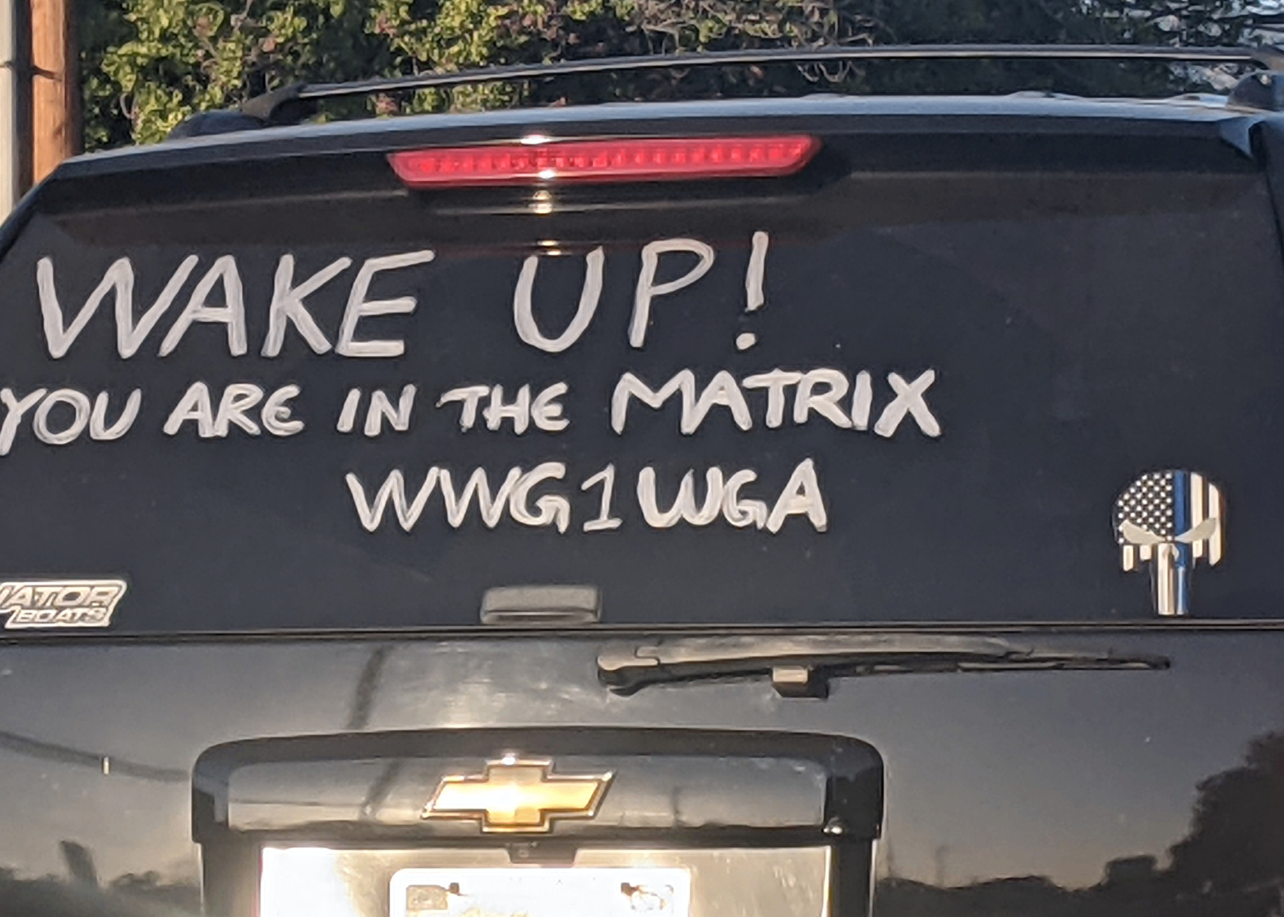 Image showing a car with the text "WAKE UP! YOU ARE IN THE MATRIX WWG1WGA" painted on the back windshield along with a thin blue line Punisher logo, all representative of the online conspiracy theory QAnon